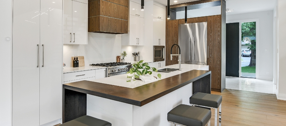 Modern kitchen with a sleek design featuring a large island with a dark wood countertop and stools, glossy white cabinets, stainless steel appliances, and a potted plant, conveying a clean and contemporary feel.
