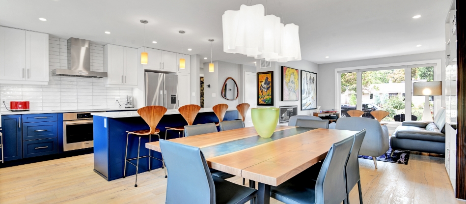 Modern open-concept kitchen and living area with blue cabinetry, wooden dining table, and vibrant art. Cozy, bright space with large windows.