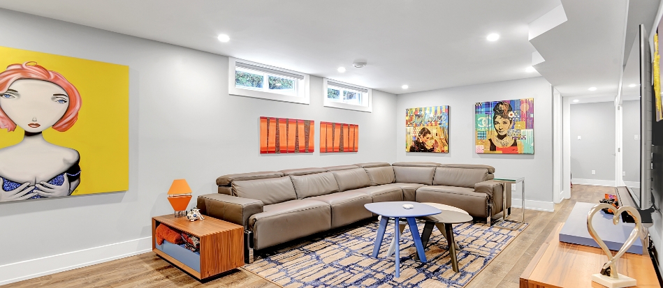 A modern living room with light gray walls features a large sectional sofa, colorful abstract art, a round table, and a blue-patterned rug, creating a vibrant and cozy atmosphere.