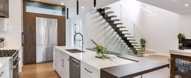 Modern kitchen with a white island, built-in sink, and greenery. Stainless steel fridge and sleek black staircase with glass railing add contemporary style.